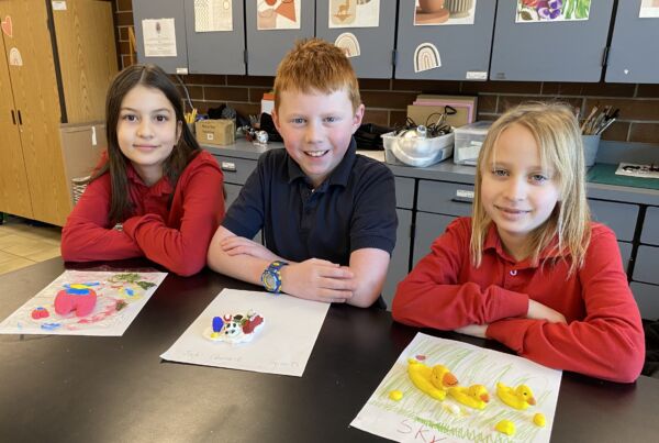 Three students with their arms folded, smiling, sitting at an art table in front of their yellow and pink artwork.