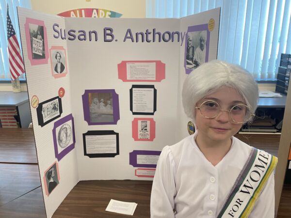 girl with a white wig standing in front of a three fold poster board. She is dressed up as, and presenting as Susan B. Anthony